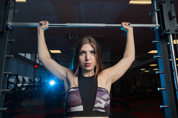 Hermosa sexy modelo de fitness sacude una barra de pesas sentado detrás de la cabeza en el gimnasio
. - Foto, imagen