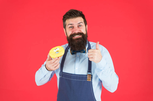 Cafe and bakery concept. Sweet donut from baker. Man bearded baker in cooking apron hold cute dessert. Ways to reduce hunger and appetite. Hipster bearded baker hold glazed donut on red background - Foto, Imagen