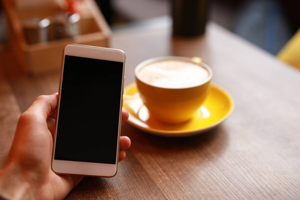 Male hand holding smartphone in cafe, yellow cup with latte on background - Фото, зображення
