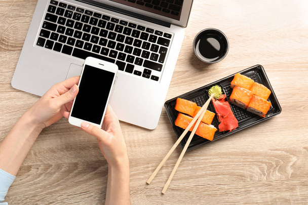 Woman with smartphone, laptop and tasty sushi rolls at wooden table, space for text. Food delivery - Foto, Imagen
