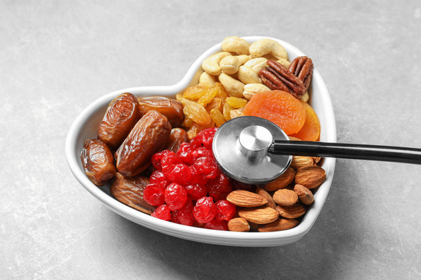 Heart shaped bowl with dried fruits, nuts and stethoscope on grey background - Фото, зображення