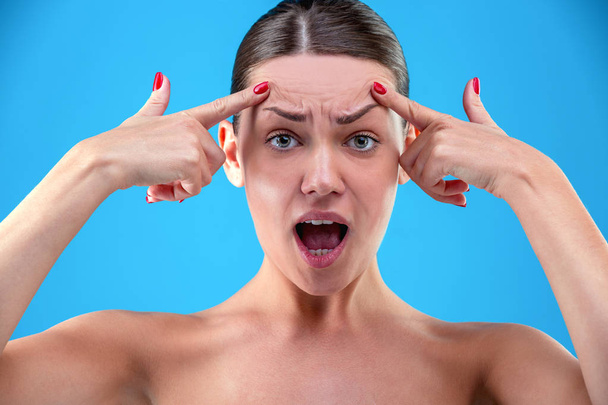 Signs of aging. Portrait of shocked young woman is looking at camera with disappointedly while touching her wrinkles on forehead on blue background - Photo, Image