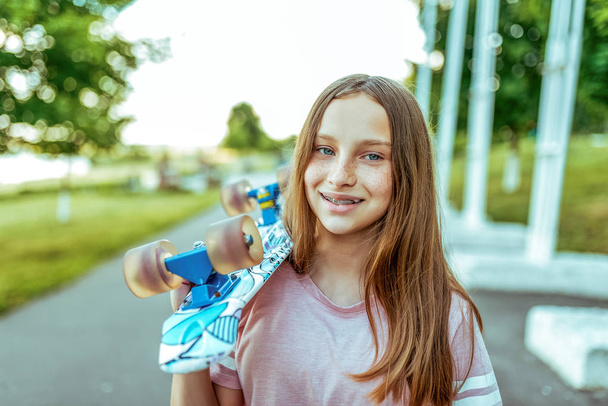 Teenager girl 10-12 happy smiling in freckles and with braces on teeth, in summer in the city, on the shoulder a skateboard for skating. Long hair, pink t-shirt. - Valokuva, kuva