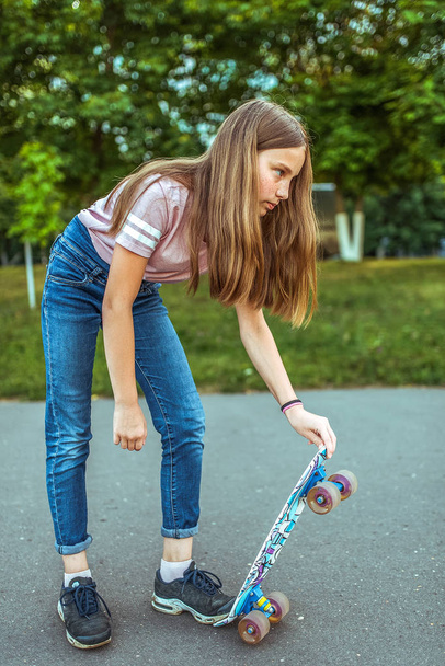Girl teenager 9-11 years old, riding a skateboard. In the summer in the city in casual jeans and sneakers. Walk in the summer park, after school lessons, rest in the city. - 写真・画像