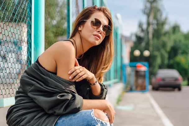 Fashion portrait of trendy young woman wearing sunglasses, jeans with halls and bomber jacket in the city - Photo, image