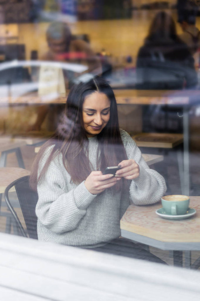 portrait beautiful smiling young woman using mobile phone in cafe throuth glass window - Фото, изображение