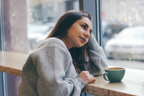 portrait of young beautiful smiling brunette girl in grey sweater drinking coffee at a table in  cafe - Photo, Image