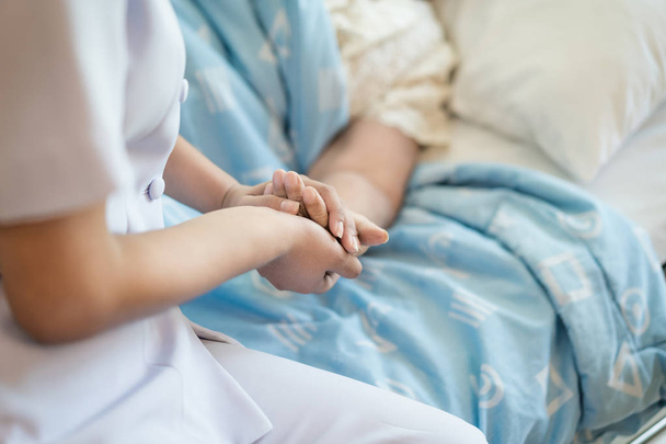 Νοσοκόμα κάθεται σε ένα κρεβάτι νοσοκομείου δίπλα σε μια ηλικιωμένη γυναίκα βοηθώντας h - Φωτογραφία, εικόνα