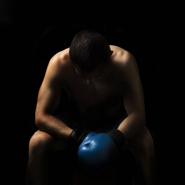A man in boxing gloves is sitting on a chair in the corner of the ring against a dark background. Boxing concept, boxing training, sparring - Фото, изображение