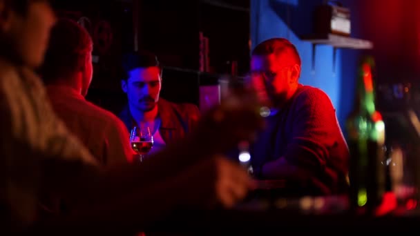 Bar with neon lighting. Company of friends drinking beer. A young man on a foreground sees his friends and sit down next to them - Footage, Video