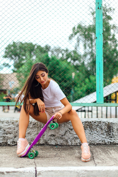Summer lifestyle image of trendy pretty young girl sitting next to the skateboard coart with her plastic skateboard. - Φωτογραφία, εικόνα