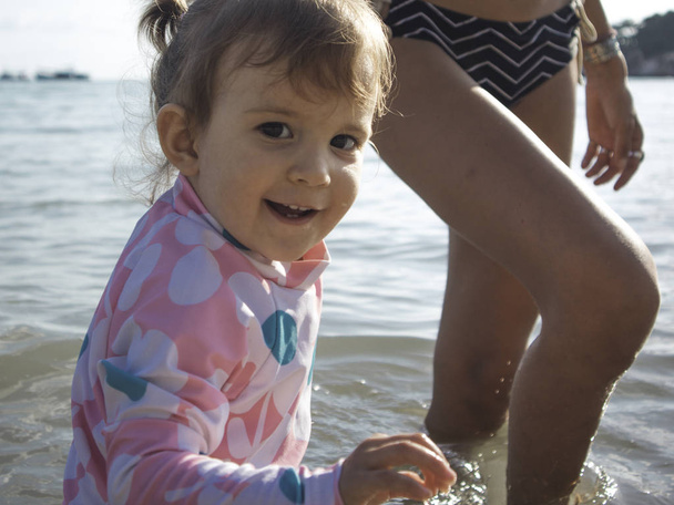 Äiti nauttii kesäpäivästä rannalla ja uima-altaalla Äiti antaa tyttärelleen reppuselässä seisoo rannalla
. - Valokuva, kuva