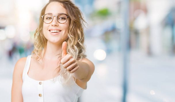 Beautiful young blonde woman wearing glasses over blurred background doing happy thumbs up gesture with hand. Approving expression looking at the camera with showing success. - Photo, Image