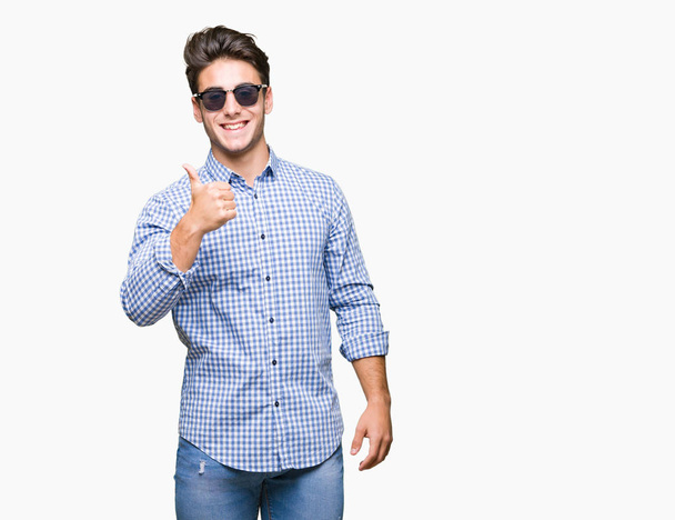 Mladý pohledný muž nosí sluneční brýle izolované pozadí dělá šťastné palec nahoru gesto rukou. Schvalování výraz s ukazuje úspěch při pohledu na fotoaparát. - Fotografie, Obrázek