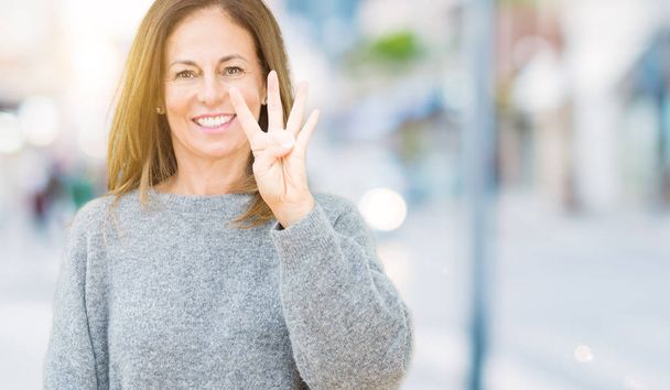 Piękne średnim wieku kobieta noszenia sweter zimowy na białym tle Wyświetlono i skierowaną w górę palcami numer cztery jednocześnie uśmiechając się pewnie i wygodnie. - Zdjęcie, obraz