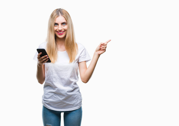 Молода красива блондинка використовує смартфон над ізольованим тлом дуже щаслива, вказуючи рукою і пальцем на бік
 - Фото, зображення