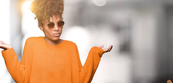 Linda jovem afro-americana vestindo óculos de sol sobre fundo isolado e expressão confusa com braços e mãos levantadas. Conceito de dúvida
. - Foto, Imagem