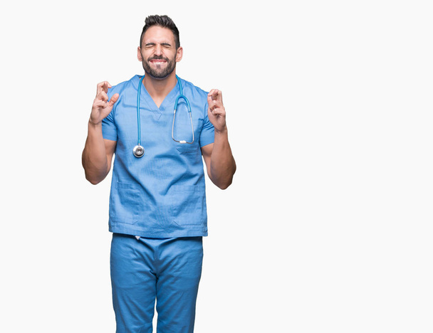 Knappe jonge dokter chirurg man over geïsoleerde achtergrond glimlachend kruising vingers met hoop en ogen gesloten. Geluk en bijgelovig concept. - Foto, afbeelding