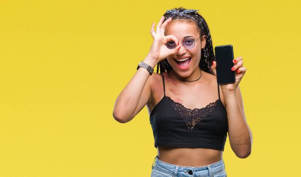 Młody pleciony włosy Afroamerykanów z znak urodzenia pokazywanie ekranu smartfona na białym tle z szczęśliwy twarz uśmiechający się robi ok znak ręką na oko patrząc przez palce - Zdjęcie, obraz