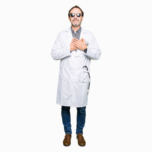 Handsome middle age doctor man wearing sunglasses smiling with hands on chest with closed eyes and grateful gesture on face. Health concept. - Foto, afbeelding