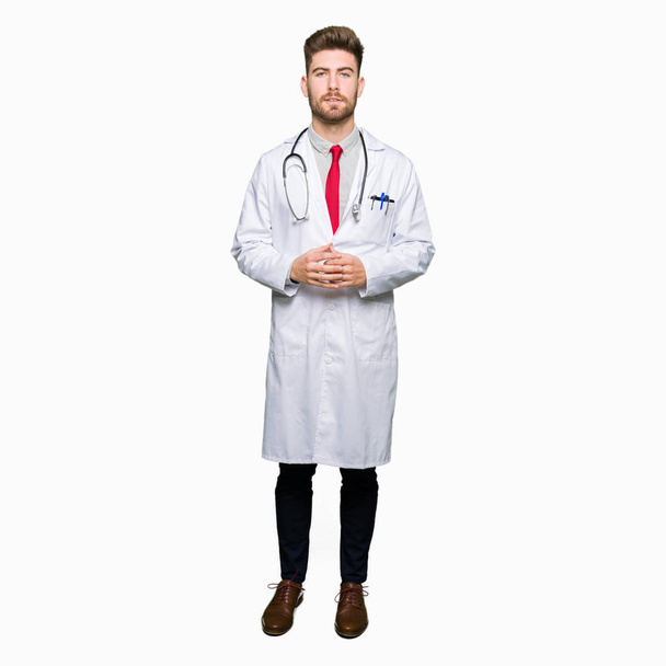 Young handsome doctor man wearing medical coat Hands together and fingers crossed smiling relaxed and cheerful. Success and optimistic - Foto, Imagen