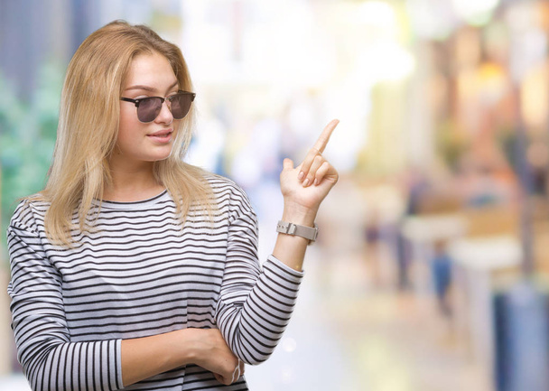 Young caucasian woman wearing sunglasses over isolated background with a big smile on face, pointing with hand and finger to the side looking at the camera. - Fotografie, Obrázek