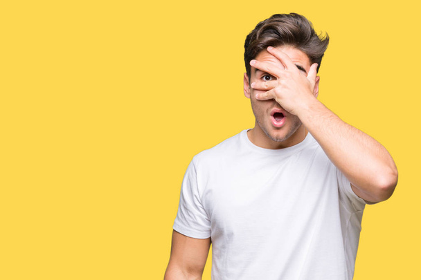 Young handsome man wearing white t-shirt over isolated background peeking in shock covering face and eyes with hand, looking through fingers with embarrassed expression. - Foto, imagen