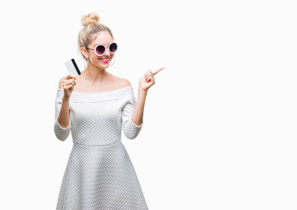 Jeune belle femme blonde tenant carte de crédit sur fond isolé très heureux pointant avec la main et le doigt sur le côté
 - Photo, image