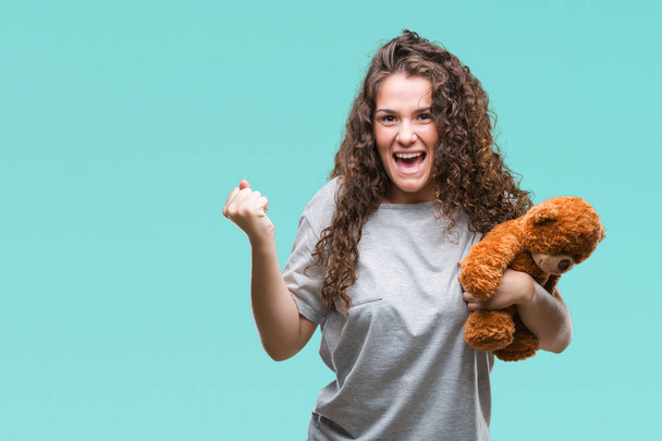 Young brunette girl holding teddy bear over isolated background screaming proud and celebrating victory and success very excited, cheering emotion - Foto, Imagen
