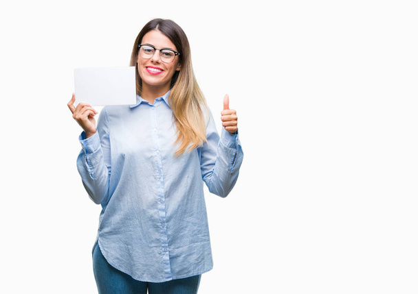 Young beautiful businesswoman holding blank card over isolated background happy with big smile doing ok sign, thumb up with fingers, excellent sign - Φωτογραφία, εικόνα