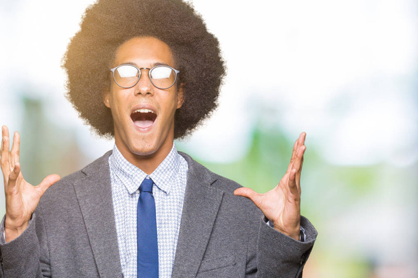 Jovem homem de negócios afro-americano com cabelo afro usando óculos loucos e loucos gritando e gritando com expressão agressiva e braços levantados. Conceito de frustração
. - Foto, Imagem