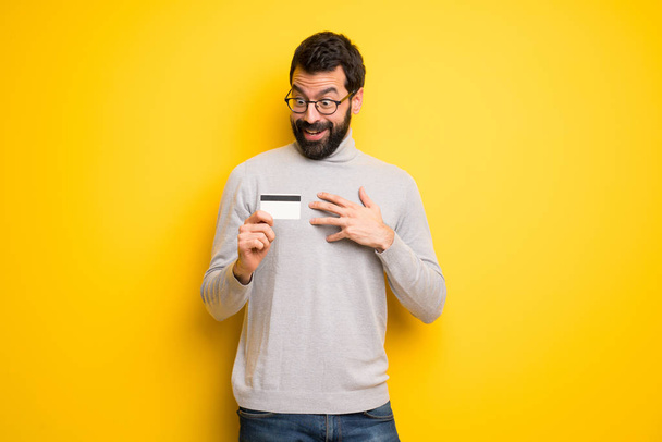 Uomo con barba e dolcevita in possesso di una carta di credito e sorpreso
 - Foto, immagini