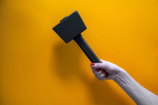 hand of a young woman holding a black wooden hammer, nails covered with red varnish, bright yellow background, close-up - Photo, Image