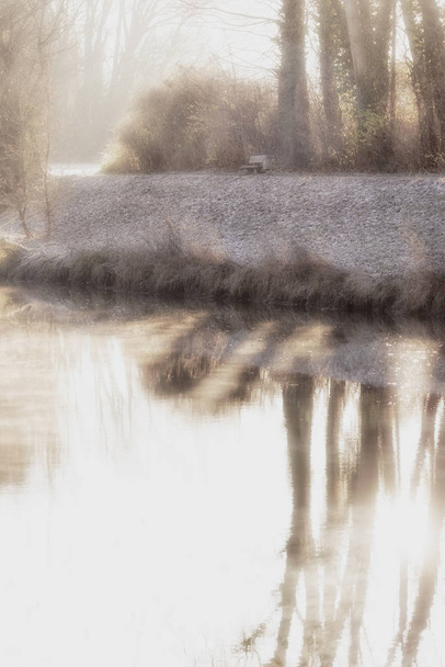 Κυκλοθυμική σουρεαλιστική ομιχλώδη φθινοπωρινό/πτώση ή χειμερινή Ανατολή στον ποταμό, αντανακλάσεις του νερού των δέντρων, παγκάκι να ξεκουραστεί στο πλάι με τα πόδια/περιπάτου/διαδρομής - Φωτογραφία, εικόνα
