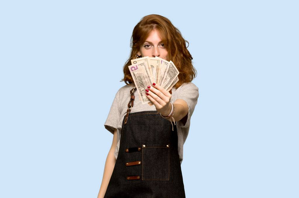 Jovem ruiva com avental tendo um monte de dinheiro sobre fundo azul
 - Foto, Imagem