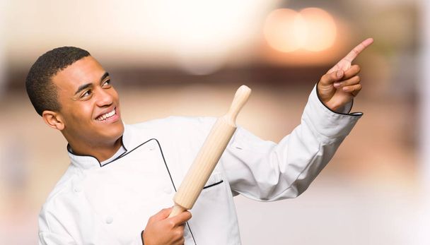Ο άνθρωπος νεαρός afro Αμερικανός σεφ δείχνοντας με το δάχτυλο δεικτών και κοιτώντας ψηλά σε αόριστες φόντο - Φωτογραφία, εικόνα