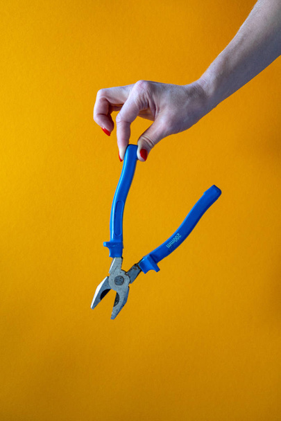 the hand of a young woman holds metal pliers with blue handles, the girl has red nail polish, close-up on a yellow background, vertical shot - Valokuva, kuva