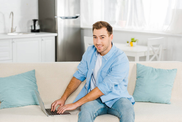 handsome man sitting on couch, looking at camera and using laptop at home - Фото, изображение