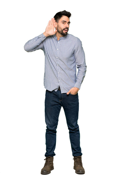 Ολόσωμο πυροβολισμό κομψό άνδρα με πουκάμισο ακούγοντας κάτι βάζοντας το χέρι στο αυτί στο απομονωμένο λευκό φόντο - Φωτογραφία, εικόνα