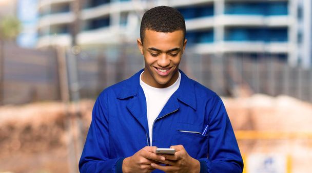 Молодой афро-американский рабочий отправляет сообщение с мобильным телефоном на строительной площадке
 - Фото, изображение