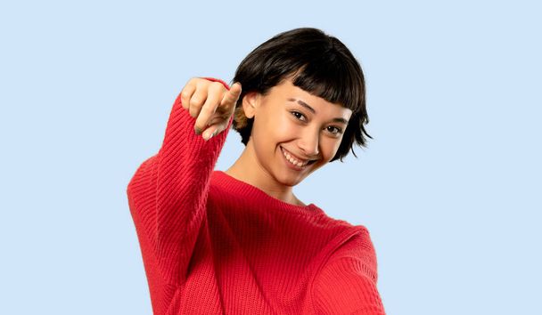 Short hair girl with red sweater points finger at you while smiling on isolated blue background - Foto, afbeelding