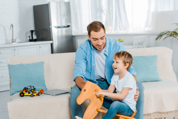 happy preschooler son riding wooden rocking horse while smiling father sitting on couch at home - Φωτογραφία, εικόνα