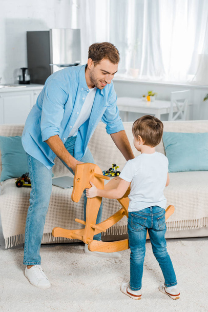 handsome father and preschooler son holding wooden rocking horse at home - Foto, Imagen