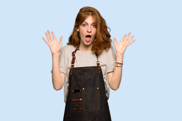 junge rothaarige Frau mit Schürze und schockiertem Gesichtsausdruck vor blauem Hintergrund - Foto, Bild