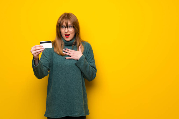 Vrouw met bril over gele muur holding per creditcard en verrast - Foto, afbeelding