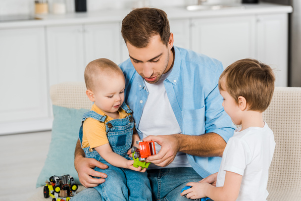 όμορφος ο πατέρας κάθεται στο καναπέ, ενώ χαριτωμένο γιους preschooler και μικρό παιδί παίζει με αυτοκινητάκια στο σπίτι - Φωτογραφία, εικόνα