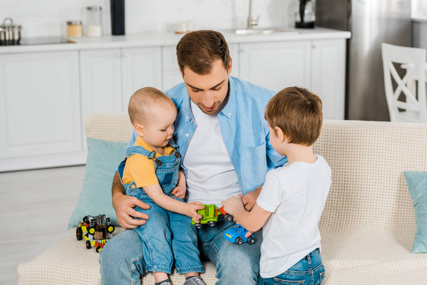 padre guapo sentado en el sofá, mientras que los niños en edad preescolar y niños pequeños jugando con coches de juguete en casa
 - Foto, Imagen