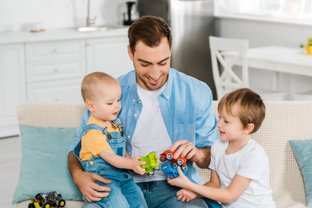 padre guapo sentado en el sofá mientras sonríe preescolar y niños pequeños jugando con coches de juguete en casa
 - Foto, Imagen