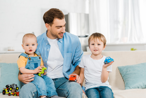 happy father sitting on couch while preschooler and toddler sons holding toy cars at home - Photo, Image