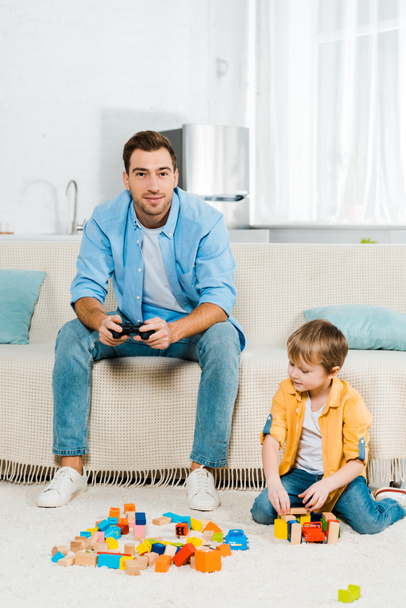 handsome father with joystick playing video game while cute preschooler son playing with colorful building blocks at home - Photo, Image